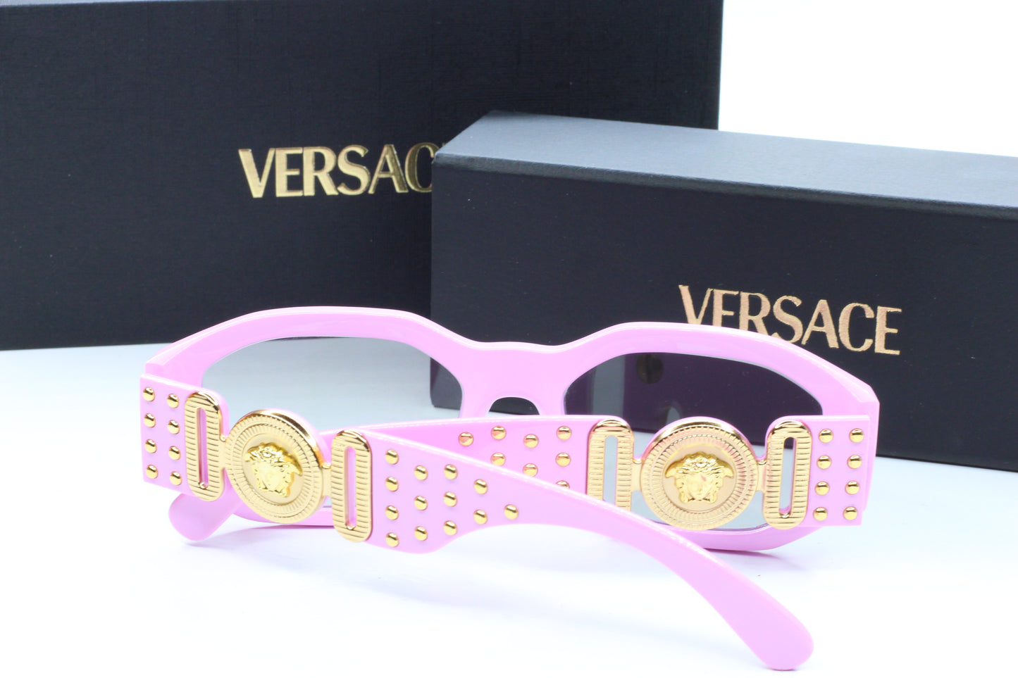 Pink Versace sunglasses with gold accents on a white surface, with Versace logo in the background.